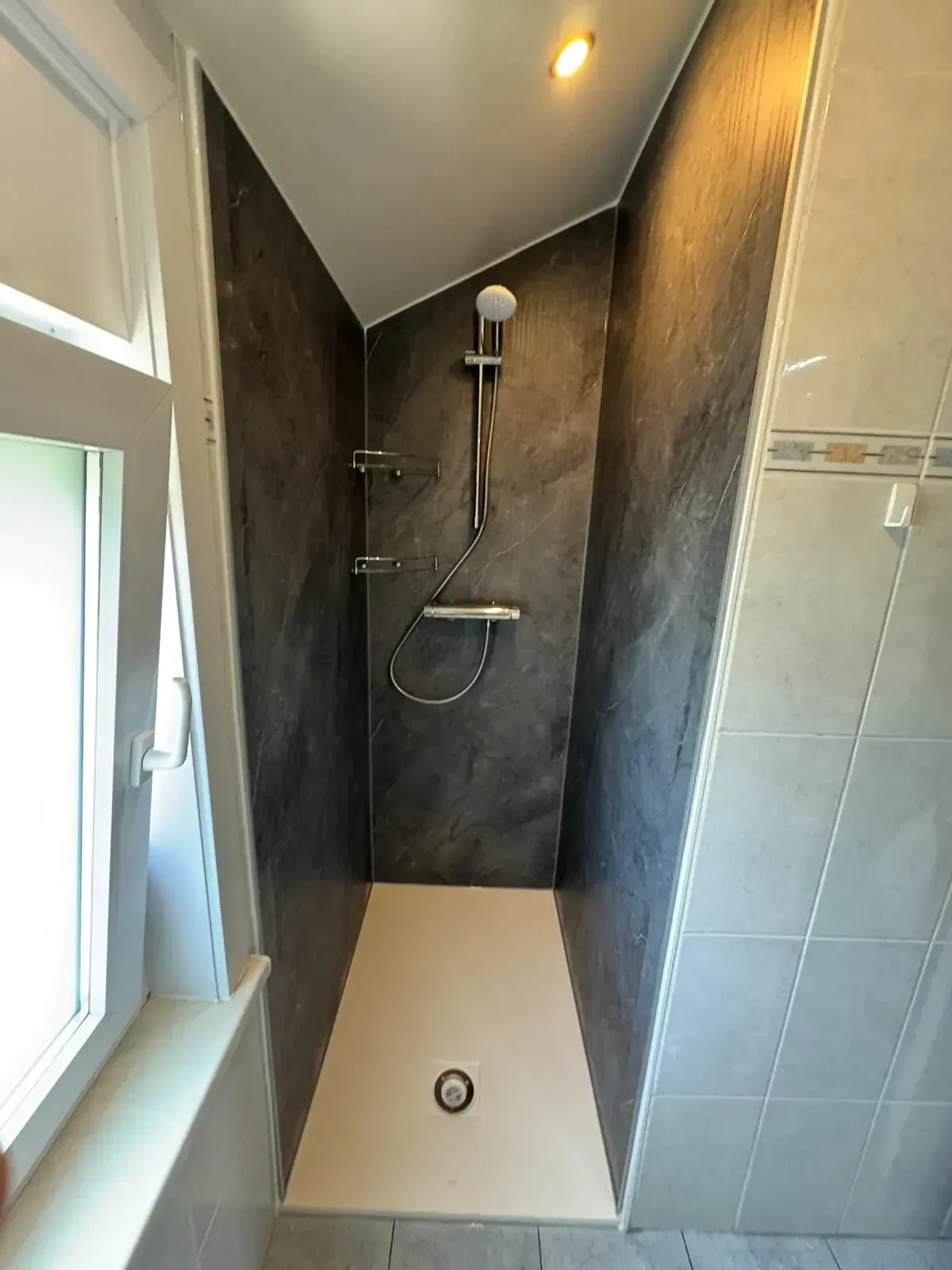 Douche onder schuin plafond met donkere marmerlook panelen, thermostaatkraan en witte douchebak bij het raam