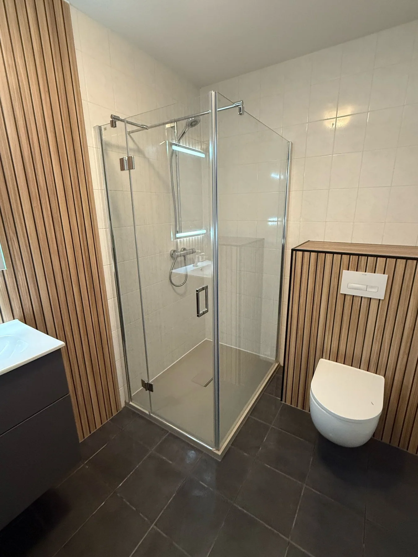 Douchecabine met glazen deur en hangtoilet tegen een voorzetwand met houtlook lamellen