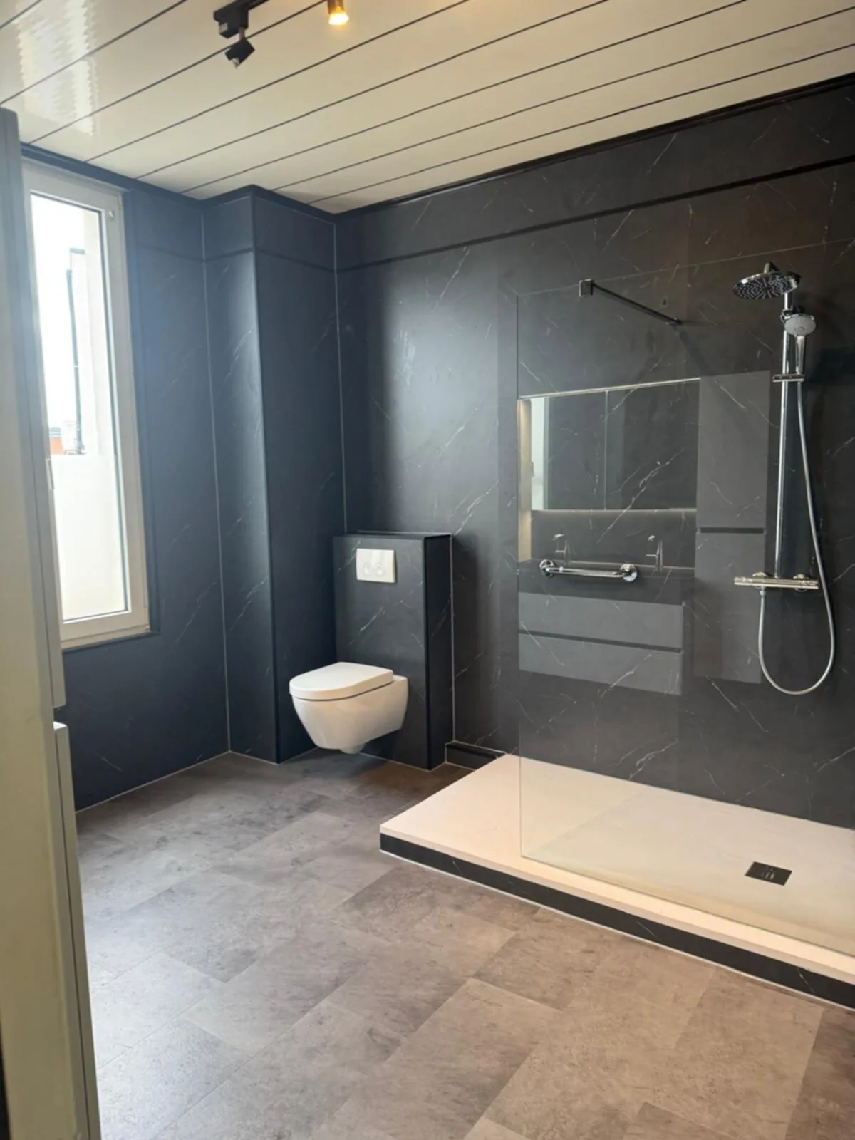 Ruime badkamer met donkere marmerlook wandpanelen, hangtoilet, inloopdouche en raam