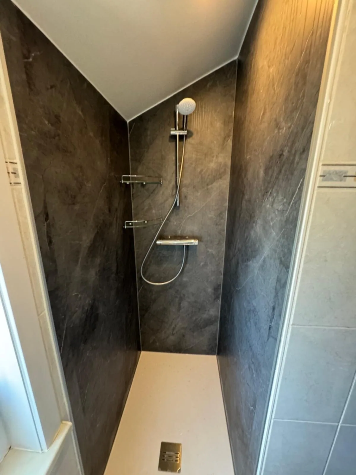 Smalle douche met donkere marmerlook wandpanelen, thermostaatkraan, planchets en witte douchebak