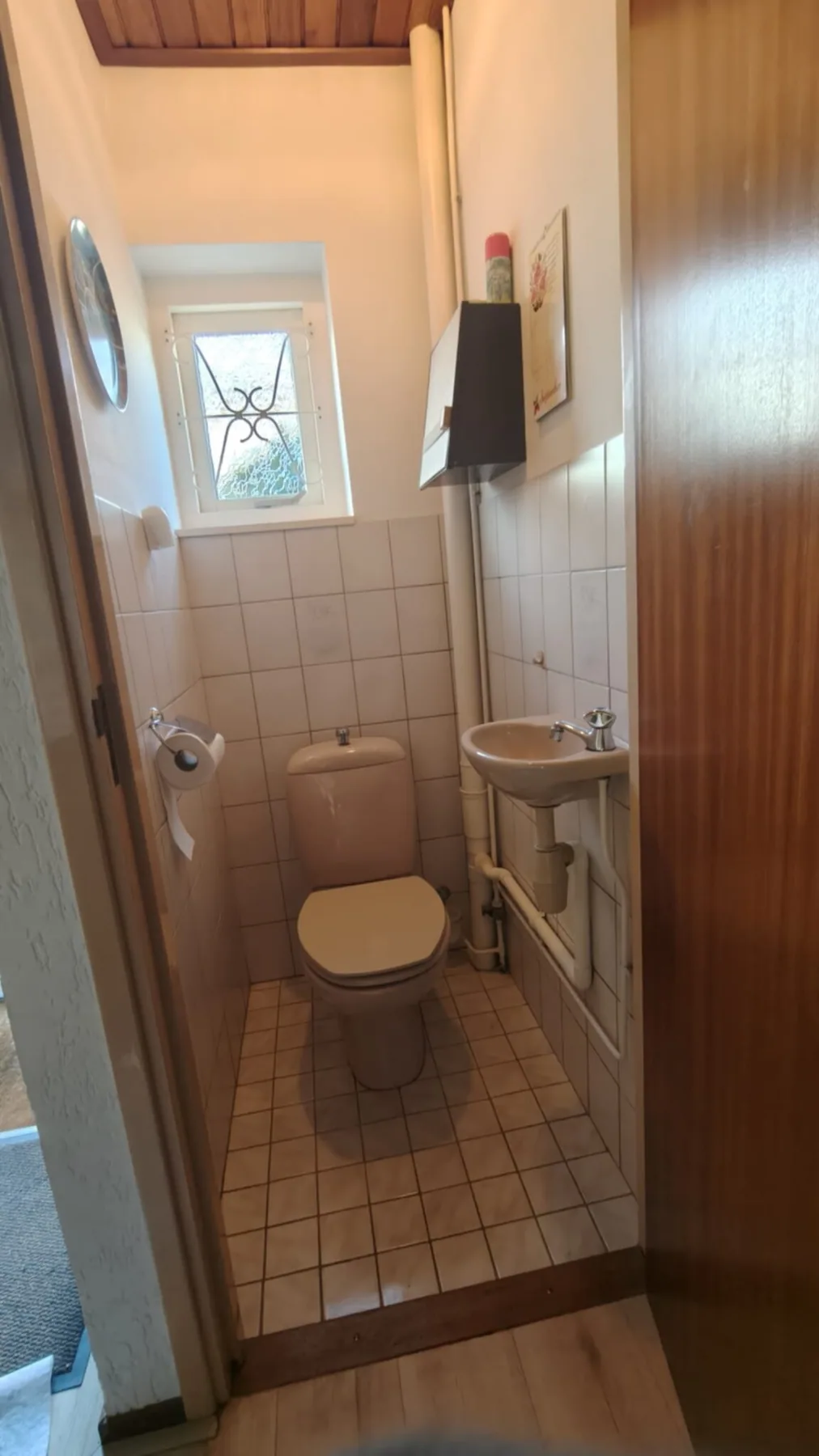 Oud toilet met beige staand closet, klein fonteintje, witte tegels en raampje met sierrooster