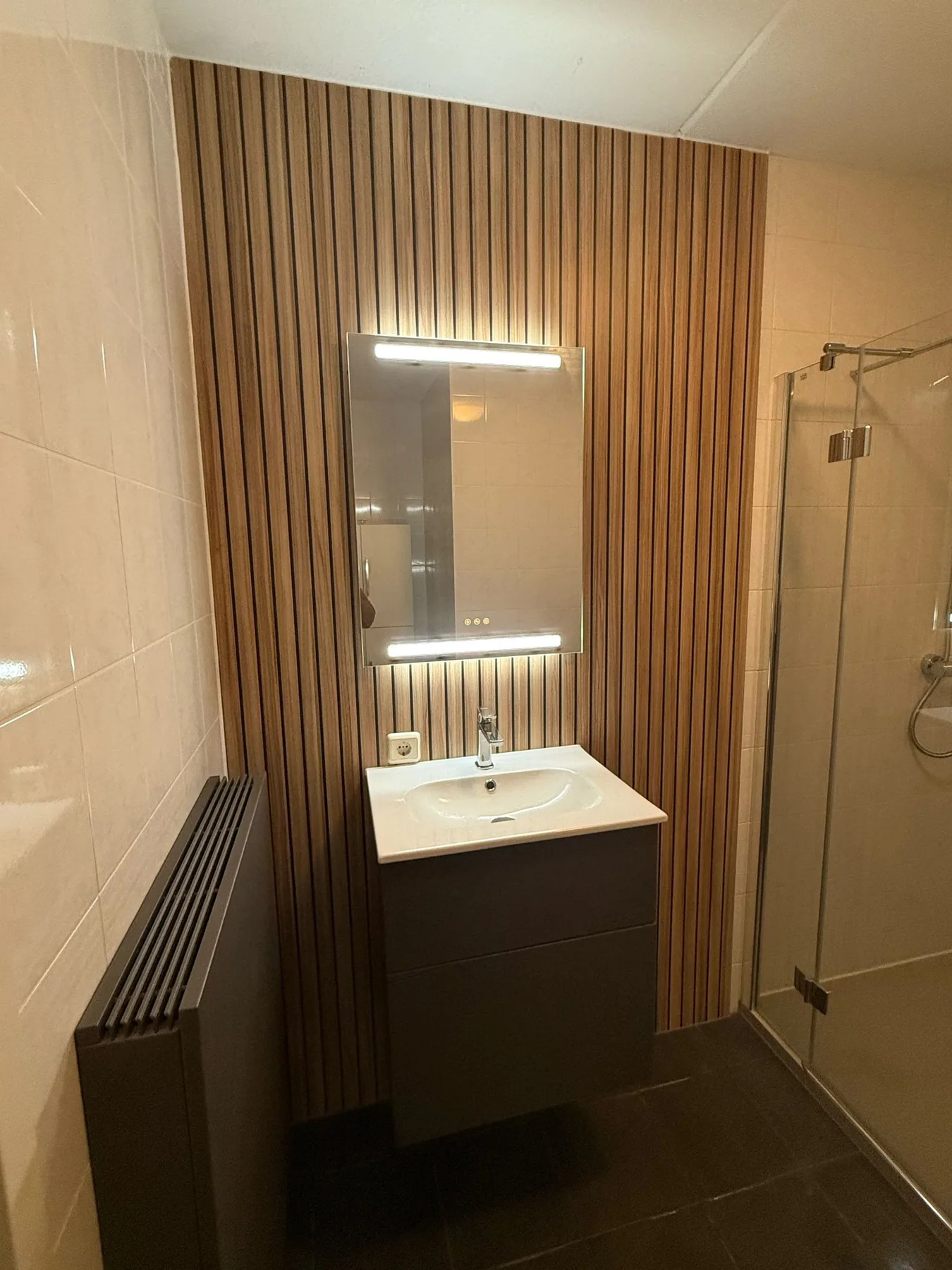 Wastafelmeubel met LED-spiegel tegen een wand met houtlook lamellen, glazen douchedeur ernaast
