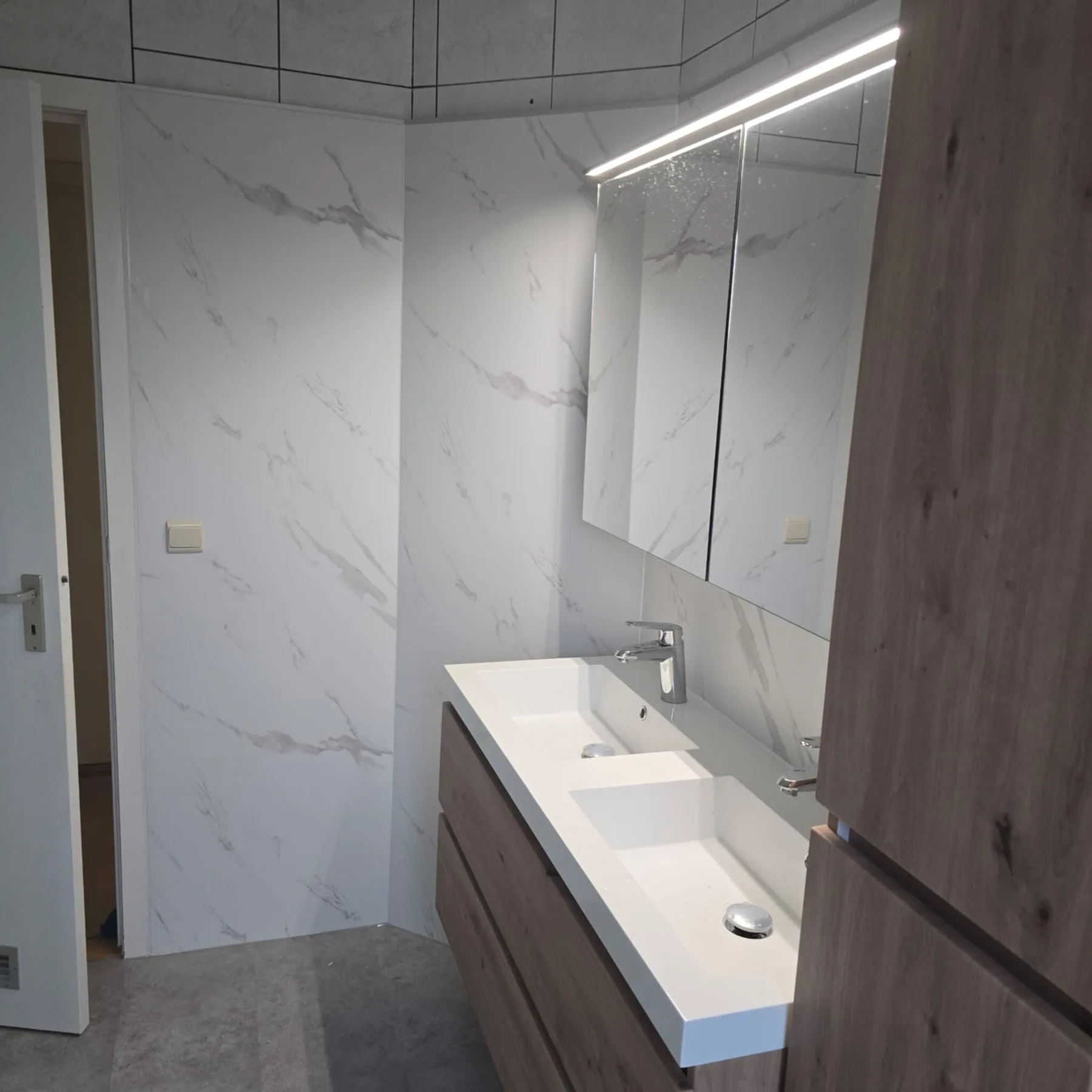 Dubbel wastafelmeubel met spiegelkast en ledbalk naast witte marmerlook wandpanelen, gezien vanaf de deur